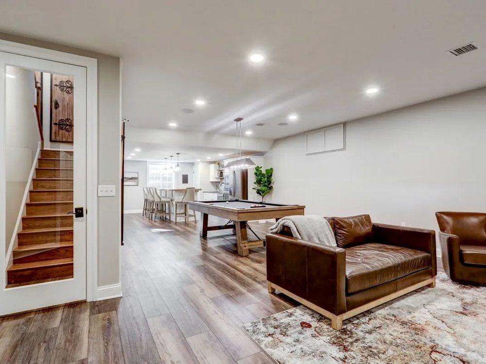 Finished basement with a seating area, games table and stairs up to the main floor