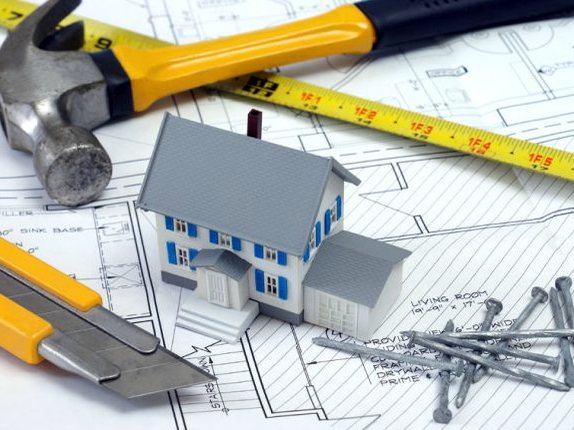 Hammer, tape measure and nails laid on a set of house blueprints beside a model home
